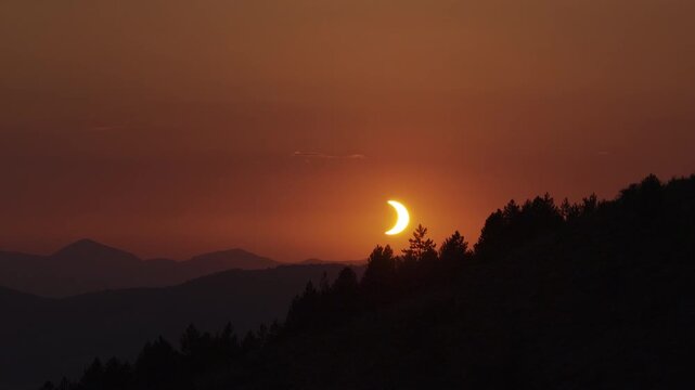 Close partial solar eclipse crescent against deep orange sky above silhouetted pine covered mountain ridge