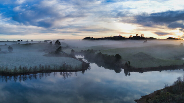 Aerial view of Lake Ratapiko at dawn with mist covering the rolling hills and calm water reflecting the sky in Ratapiko, Taranaki Region, New Zealand.