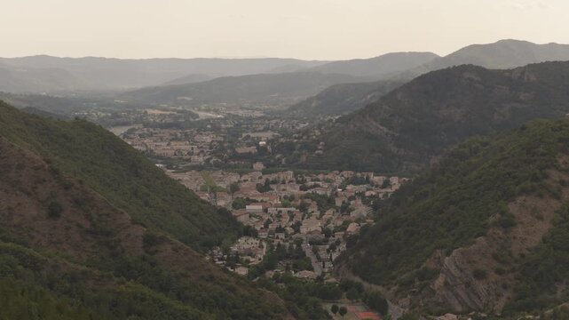 High overlook of dense mountain town in narrow green valley with surrounding forested ridges and distant peaks