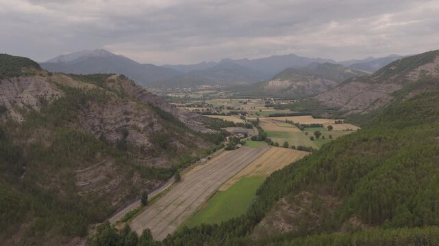 Wide aerial of green valley agricultural fields road and gray eroded mountain slopes under cloudy sky