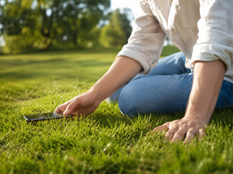 Person sitting on green grass outdoors reaching toward the ground beside a smartphone while taking a mindful screen break to reconnect with nature and find calm balance stress relief