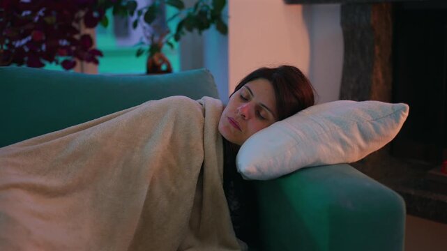 Adult woman naps beneath a blanket on couch at home with her head resting on a pillow, expressing fatigue, peaceful recovery, comfort
