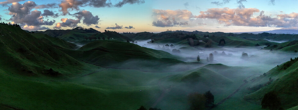 Aerial view of the rolling green hills of Tarata blanketed in morning mist under a soft sunrise sky in Tarata, Taranaki Region, New Zealand.