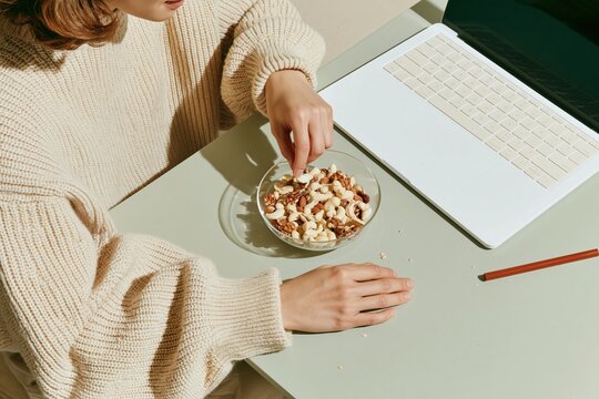 East Asian woman reaching for mixed nuts in a glass bowl on a home-office desk, featuring cozy knit textures for healthy snacking and remote work