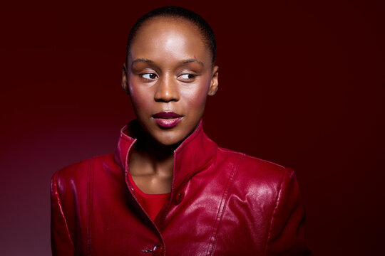 Portrait of a young black woman with dark skin wearing a red leather jacket, looking sideways against a red background. Concept of fashion, confidence, and beauty in studio lighting.