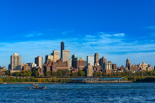 The scenic urban skyline of Brooklyn, New York features modern high-rises and historic buildings viewed from across the river with a small boat cruising on the water.
