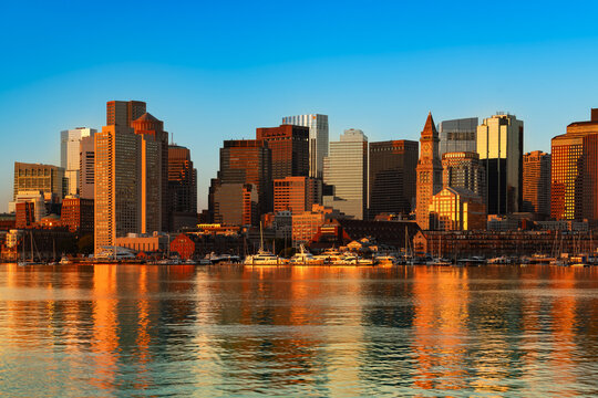 Golden morning sunlight illuminates the downtown Boston skyline across the peaceful bay at sunrise, with skyscrapers reflecting warm light on the calm harbor water.