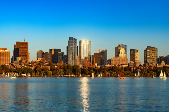 Sunlight reflects off modern glass skyscrapers of Back Bay and the Beacon Hill area in Boston as sailboats glide along the calm waters of the Charles River.