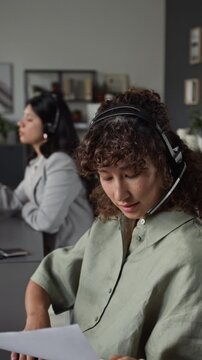 Young woman wearing headset working in call center, talking to client and reading document. Clip is suitable for customer service, telemarketing, and corporate support, vertical