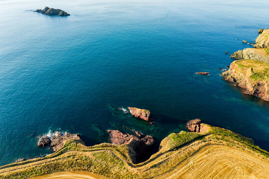 Elevated view of coastal cliff top with winding walking trail, rocky outcrops and reefs visible through turquoise shallows beside deep ocean water