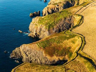 Winding footpath along grassy cliff edge in Pembrokeshire, Wales, evening sunlight illuminating...