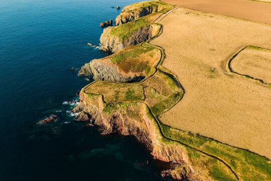 Winding coastal footpath along steep rocky headlands bordering dry pasture fields, warm evening light over the coastline in South Wales