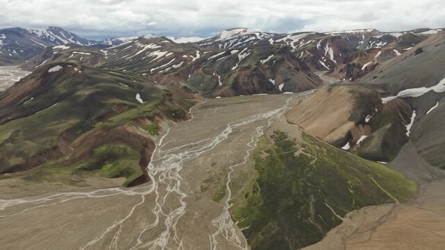 A stunning aerial push in shot of the Reykjakollur mountain near Laugavegur hiking trail on Iceland. Showcases the rugged, colorful landscape with braided rivers and snow patches under a cloudy sky.