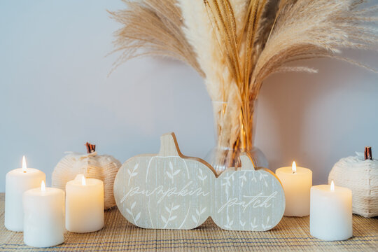 Cozy warm autumn home interior - white knitted pumpkins, wooden statuette, candles, dried flowers on the wooden console with white wall background. Scandinavian minimalist hygge home fall decor.