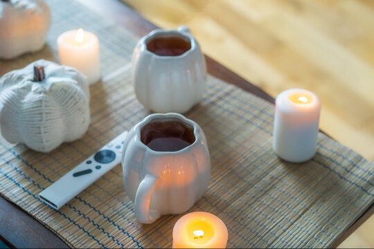 Autumn fall mood composition for hygge home decor. White pumpkin's shaped cups with hot drink, burning candles, remote controller on coffee table in living room. Movie night at home. Cozy relax time