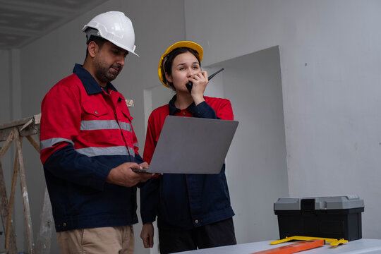 Construction engineers coordinating with a two-way radio, reviewing project data on a laptop at an indoor building site, emphasizing teamwork, site communication, project management, quality control.