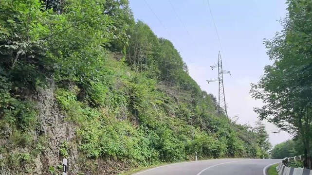 Driving on winding mountain road through lush green forest with electricity pylon, scenic travel adventure along steep cliffside on beautiful summer day with clear sky