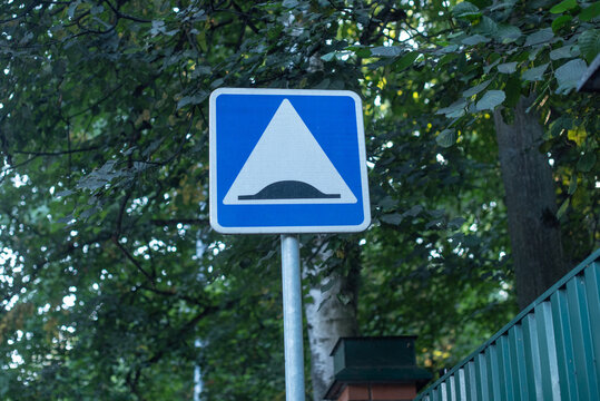 A blue triangular road sign indicating a speed bump, surrounded by green trees and foliage. The sign is mounted on a metal pole.