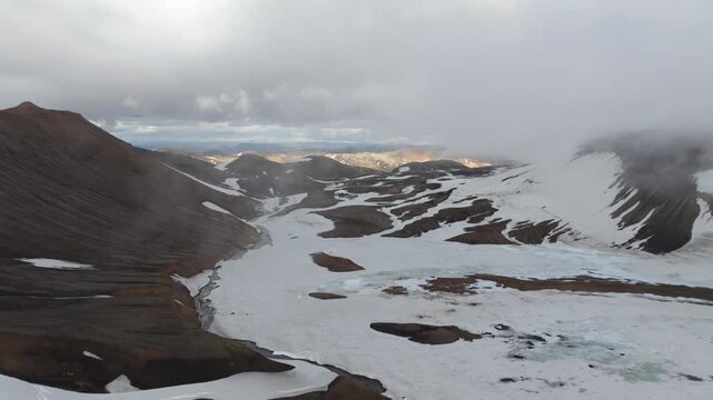 Stunning aerial pull back along St&ouml;&eth;ull mountain and the Laugavegur trail on Iceland. The vast, rugged landscape features a glacial river winding through snowfields and rocky hills.