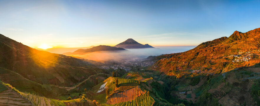 Aerial view of Sikapuk Hill and Villa Dieng at sunrise with terraced agricultural fields and a distant volcano rising above the morning mist in Indonesia.