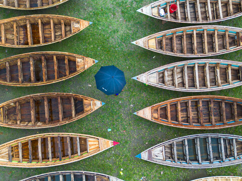 Aerial view of wooden boats arranged in rows on a grassy field with a person holding a dark umbrella in the center in Manikganj, Dhaka Division, Bangladesh.