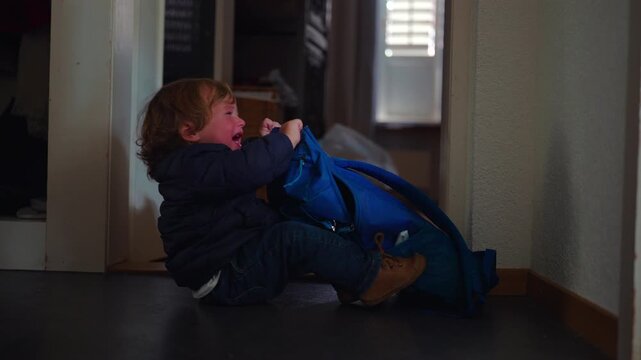 Crying toddler throws a tantrum while pulling a blue backpack on the home hallway floor, expressing frustration, emotional overload and early childhood independence