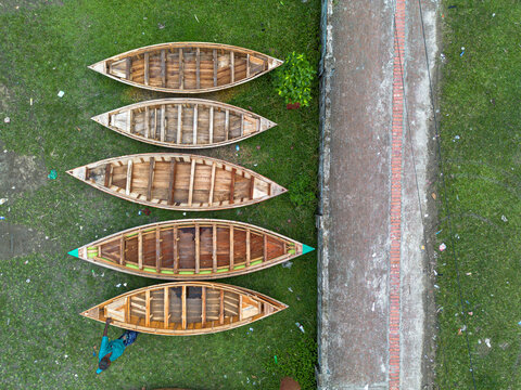 Aerial view of wooden boats lined up on the green grass next to a stone path with a man standing nearby in Manikganj, Dhaka Division, Bangladesh.