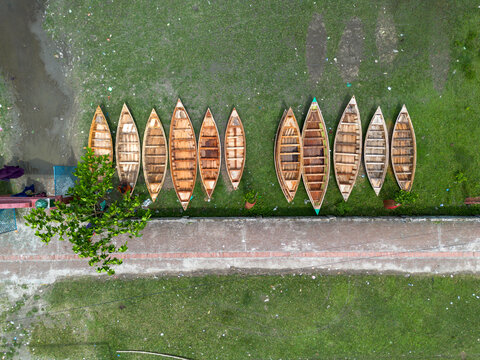 Aerial view of wooden boats lined up on a grassy bank next to a paved path and a tree in Manikganj, Dhaka Division, Bangladesh.