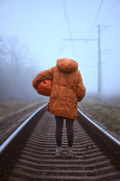 Back view of a pumpkin headed figure walking along railway tracks into the fog. Halloween concept symbolizing mystery, journey and loneliness in a misty landscape
