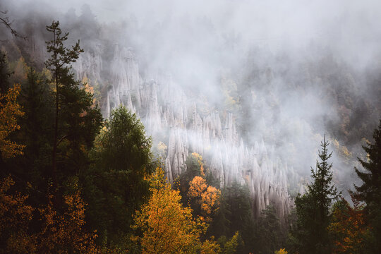 Autumn forest covered by drifting fog beneath the earth pyramids of South Tyrol. Atmospheric landscape with colorful trees and misty woodland