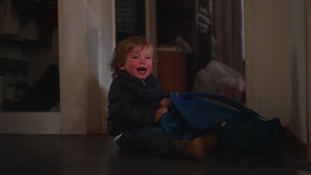 Upset young child screams while wrestling with a blue bag on the corridor floor, revealing childhood frustration, emotional overload and everyday parenting challenges