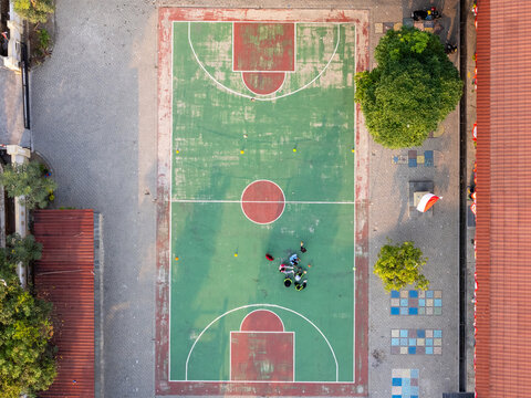Aerial view of a green basketball court with red markings and a group of people gathered in the center, surrounded by trees and tiled ground in Surakarta, Central Java, Indonesia.