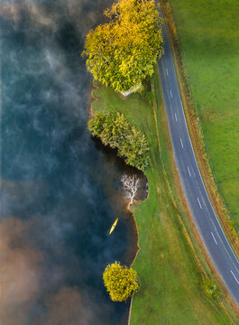 Aerial view of Lake Ratapiko with a yellow kayak on misty water, surrounded by green fields and trees next to a road in Ratapiko, Taranaki Region, New Zealand.