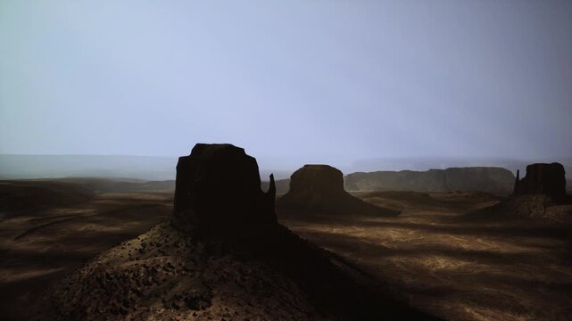Desert buttes under hazy dawn light, long shadows stretching across cracked plain, windswept sandstone monoliths rising from arid plateau, muted bluegray sky,