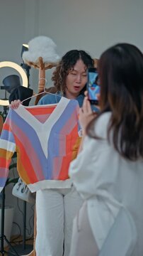 Young Asian woman fashion entrepreneur live streaming selling clothes online with smartphone ring light and laptop in home studio, representing small business owner and professional women.