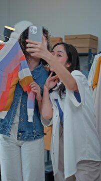 Young Asian woman fashion entrepreneur live streaming selling clothes online with smartphone ring light and laptop in home studio, representing small business owner and professional women.