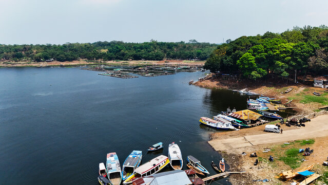 Aerial view of Jangari reservoir with numerous small boats docked at the shore and floating fish farms in the distance, surrounded by lush greenery in Cianjur, West Java, Indonesia.