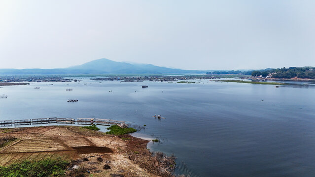 Aerial view of Jangari reservoir featuring floating fish farms, calm water, distant mountains, and agricultural land in the foreground in Cianjur, West Java, Indonesia.