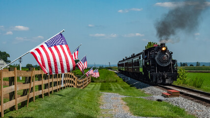 A steam train moves along the tracks surrounded by green grass and a wooden fence. American flags wave in the breeze on a sunny day in a rural area. © Greg Kelton