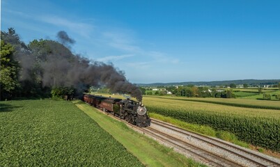 A steam train moves along tracks surrounded by green fields in a rural area. Smoke rises from the engine as it passes by a small town. The sky is clear with no clouds. © Greg Kelton