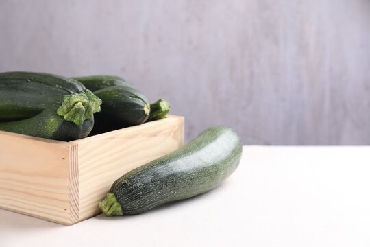 Fresh raw zucchinis in wooden crate on light table against gray background, closeup. Space for text