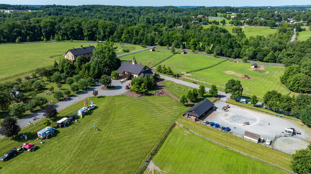 A farm area is visible with buildings scattered around. There are vehicles parked along the road. Green fields stretch in every direction. Trees and sunny sky complete the scene.