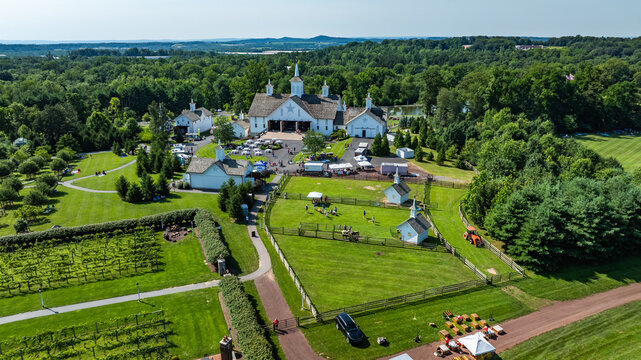 People gather at a large property with green fields and trees. Tents are set up for an outdoor event. Animals are seen in fenced areas. The sun shines on this busy summer day.