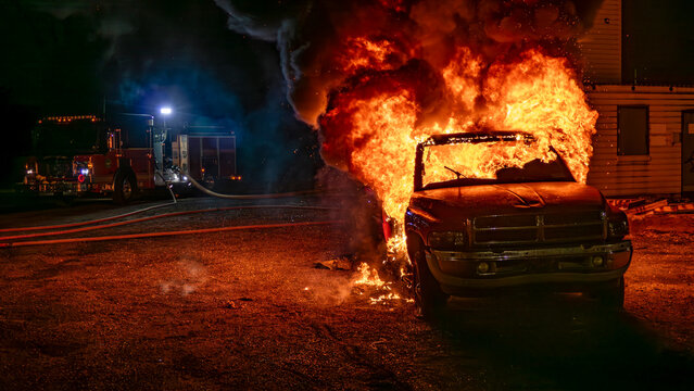Flames erupt from a car in a parking lot at night. Firefighters use hoses to control the fire as smoke billows. The area is lit by emergency lights, creating an intense scene of action.