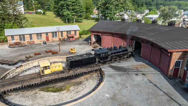 A steam train departs from a roundhouse in a small town. The sun shines bright as the train moves on tracks. Trees and buildings are seen in the background. It is a clear afternoon.