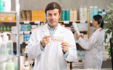 Fototapeta premium Polite middle-aged male pharmacist offering pills box while woman working in background in drugstore