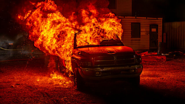 Flames rise from a truck in a training session for firefighters. Emergency responders work at night to manage the situation. Smoke billows as they practice fire control techniques.