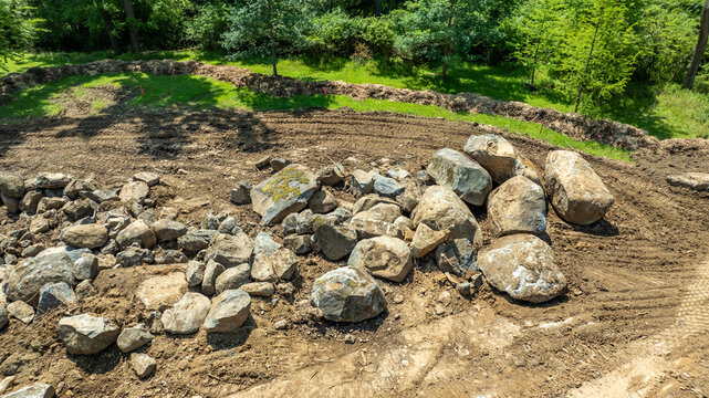 Workers move large rocks and clear soil in an outdoor area. The setting is sunny with trees nearby. Equipment may be used to prepare the ground for a construction site or landscaping project.