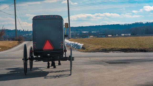An Amish horse-drawn buggy moves along a rural road surrounded by open fields and trees. The sun shines brightly and clouds are visible in the sky. This scene shows a typical day in the countryside.