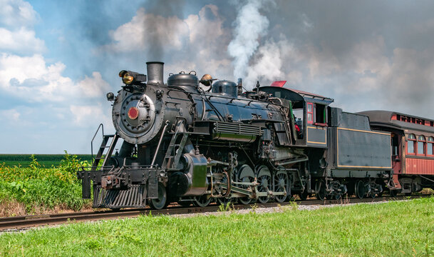 A steam locomotive moves on tracks as smoke rises into the sky. The setting is a rural area with green fields. The scene captures the power and movement of the train in daylight.